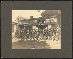 Gelatin silver print of a large group of men on bicycles in front of Albert E. McDaniel Bicycle Doctor, matted on dark gray board. A sticker on the bottom left corner of the back of the matting board reads "Mrs. Allen G. Shiek 101 Myrtle Avenue Claymont, Del. 19703".