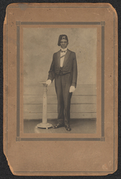 Photograph of a man standing and leaning slightly on a small pillar. He is wearing a three piece suit, white gloves, and a fez with a tassel. The photograph is in a brown card frame with black accents. The back of the frame identifies the man as "Mr. Jerry Blacke" in pen. The studio and photographer are not noted.