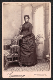 Photograph of a woman wearing a layered dress posing with a chair. The name and address of the photography studio is printed along the bottom of the card. It reads: "Cummings 302 Market Street, Wilmington, Del.".