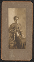 Photograph of a woman wearing a black dress sitting sideways on a chair. Mounted on a brown-gray board. Embossed under the photograph is "A.N. Sanborn 404 Market St. Wilmington, Del.".