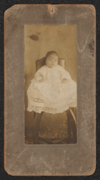 Photograph of a baby wearing a lace dress and sitting on a chair. Mounted on a gray board. Embossed beneath the photograph is "A.N. Sanborn 404 Market St. Wilmington, Del.".