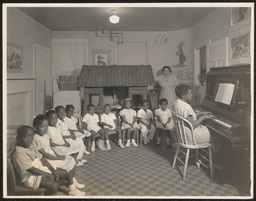Several children sit in a semi-circle around teacher at piano, with another teacher in attendance.