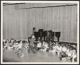 Child conducts group of children with musical instruments accompanied by teacher on piano, circa 1945-1965