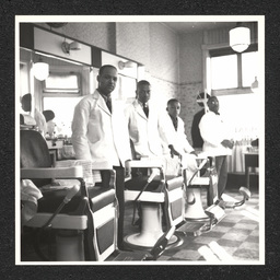 Four barber pose by their chairs in a barbershop