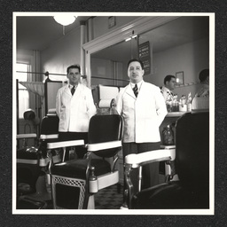 Two barbers pose by their chairs inside of a barbershop