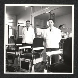 Two barbers pose by their chairs inside of a barbershop