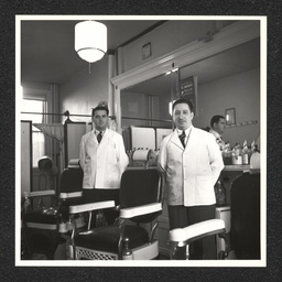 Two barbers pose by their chairs inside of a barbershop