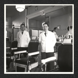 Two barbers pose by their chairs inside of a barbershop