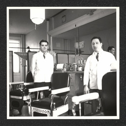 Two barbers pose by their chairs inside of a barbershop