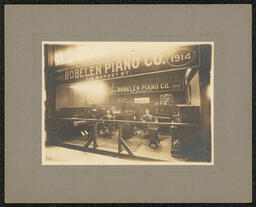 Gelatin silver print of two men in a booth surrounded by pianos, a sign that reads "Robelen Piano Company" hangs over them. The photograph was possibly taken at a trade fair. Mounted on gray board. Written on the back in pen: "W.E. Holland on right Mr. Calhoun on left".