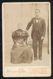 Carte de Visite of a woman and man, both wearing watches. The woman is seated, the man stands next to her with his hand on her shoulder. Information about the photography studio is printed on the lower left corner.