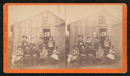 Stereoscope Card of a group of girls sitting in front of a wooden building.