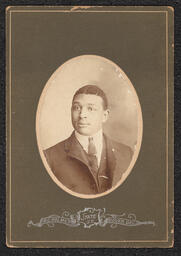 Photograph of a man wearing a suit, including a tie pin and clip. The photograph is in a dark gray frame with an oval cut out for the image. Below the image is a metallic stamp with the name and address of the photography studio: "R.C. Holmes, State St., Dover, Del.".