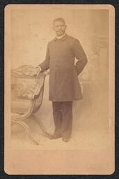 Photograph of a standing man wearing a long coat leaning against a chair. The name of the photography studio appears to be stamped on the bottom edge of the card, however the only word that can be made out is "Wilmington".