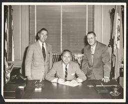 Three men in suits behind desk, 1955