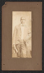 Photograph of a man wearing a suit standing and leaning on a chair. In his hand he holds a bound book. The photograph is mounted on a dark brown board. Very faintly embossed under the photograph is "Wilmington Photo Co.".
