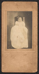 Photograph of a baby in a long white dress sitting in a chair. Mounted on a tan board. Embossed beneath the photograph is "A.N. Sanborn 404 Market Street Wilmington, Del.".