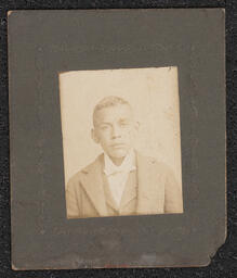 Photograph of a young boy wearing a suit jacket, taken from the shoulders up. Mounted on a dark gray board with embossed decorations around the photograph.