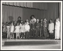 Group of smaller children performing on stage with two teachers chaperoning.