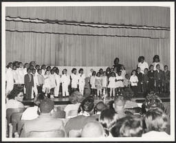 Several groups of children perform on stage with some teachers for a live audience, presumably parents and guardians.