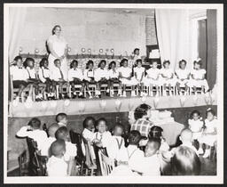 Children sit on stage in a row with a teacher standing and another teacher on piano behind them. Other students sit with a teacher in the audience.