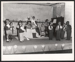 Children dance on stage in front of two other children in monarch costumes on thrones, accompanied on piano by a teacher.