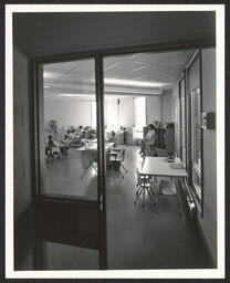View of a classroom through an open doorway. Children sit at desks in groups while teacher supervises from a chair at the front of the room.