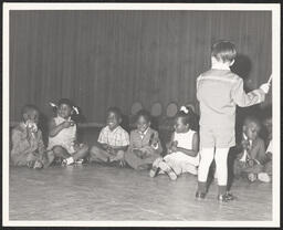 Child conducts group of seated children playing musical instruments, circa 1945-1965